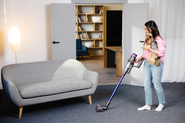 Woman with vacuum cleaner and dog in her hand vacuuming in the living room.