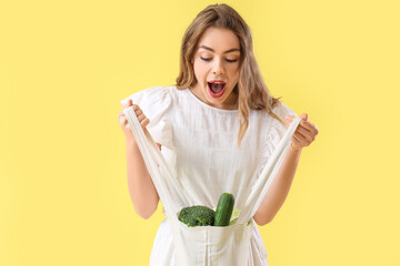 Surprised young woman with fresh vegetables in shopper bag on yellow background