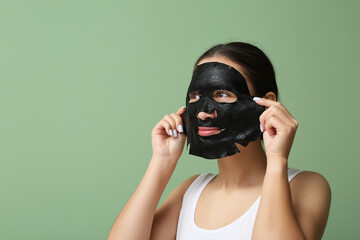 Young woman applying activated charcoal mask on her face against green background