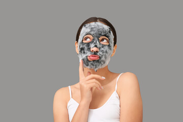 Thoughtful young woman with activated charcoal mask on her face against grey background