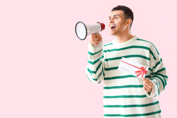Young man with gift card shouting into megaphone on pink background