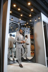 Fototapeta na wymiar sale, shopping, fashion, style and people concept - african man choosing and trying jacket or suit on and looking to mirror in mall or clothing store