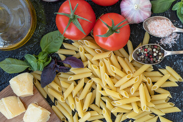 close-up top view of penne pasta, red ripe tomatoes, parmesan cheese, olive oil, fresh basil, garlic and spices. flat lay on black textured background