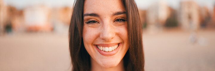 Fototapeta premium Beautiful brown-haired woman with long hair sits on the beach, Panorama. Gorgeous girl with freckles and long eyelashes looks at the camera and smilig.