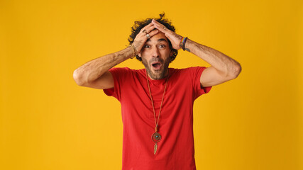 Wonder attractive man with curly hair, dressed in red T-shirt,  put his hands on his head in surprise looking at camera isolated on yellow background in studio