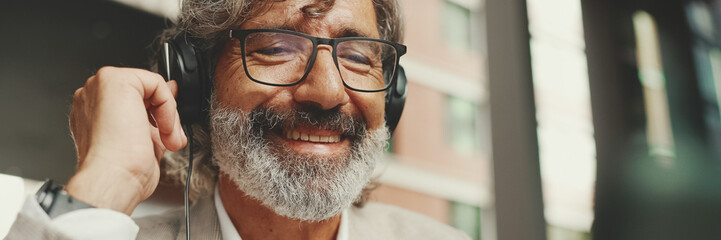 Panorama, Close up portrait of mature businessman with beard in eyeglasses and headphones, sits in...