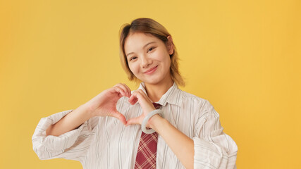 Smiling young woman wearing shirt and tie showing representing heart in shape of fingers gesture isolated on yellow background in studio