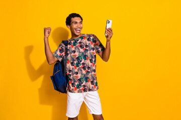 Photo of young lucky guy in t shirt raised fist up wear backpack using smartphone finally done session isolated on yellow color background