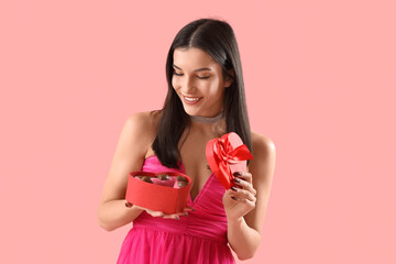 Young woman with box of heart-shaped chocolate candies on pink background