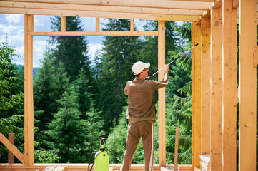 Laborer constructing wooden frame house near forest. Back view of man treating woods, applying fire retardant using sprayer. Concept of modern eco-friendly construction. - obrazy, fototapety, plakaty