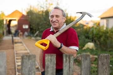 Portrait of satisfied elderly farmer with shovel near wooden fence of his farm