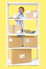 Young online store seller and rack with parcel boxes on yellow background