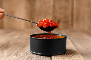 Red salmon caviar in a black tin can and a black spoon full of caviar on a wooden background.