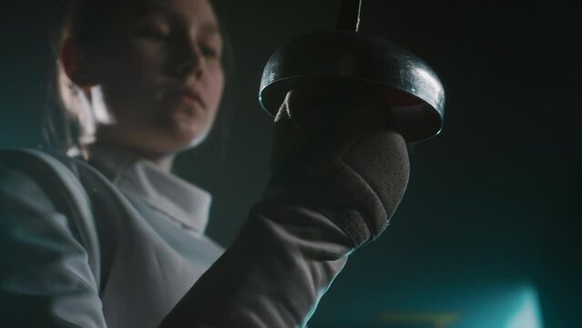 Low angle closeup with focus on rapier and female foil fencer looking at her weapon in dark hall