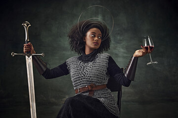 African woman, medieval warrior in chainmail armor sitting on chair with sword and wine glass over vintage green background. Brave woman. Concept of comparison of eras, history, creative art, remake - 763275531