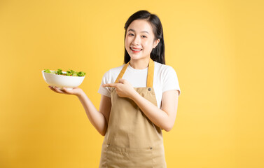 Portrait of Asian female housewife holding a bowl of salad and posing against a yellow background