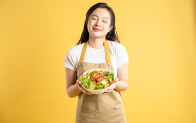 Portrait of Asian female housewife holding a bowl of salad and posing against a yellow background