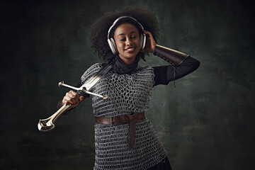 Smiling young African woman, medieval warrior in chainmail armor with sword listening to music in headphones against vintage green background. Concept of comparison of eras, history, modernity, remake