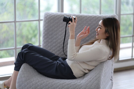Asian woman lying on sofa holding DSLR camera, reviewing photos from camera before sending photos for sale in apartment