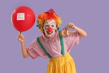 Funny little girl dressed as clown with balloon and paper fish on lilac background. April Fools' Day celebration