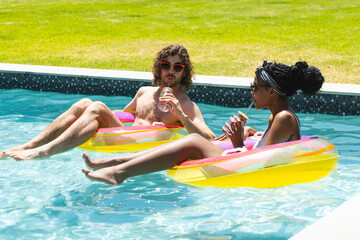 A diverse couple relaxes on pool floats, enjoying drinks at home