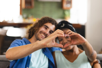 A diverse couple forms a heart shape with their hands at home on the couch