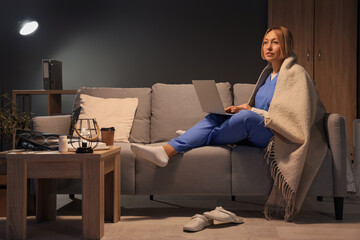 Fototapeta premium Female nurse with plaid using laptop on sofa at hospital in evening