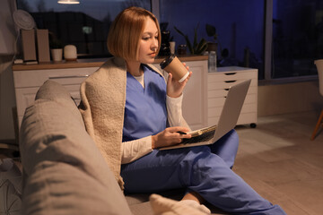 Female nurse with coffee using laptop on sofa at hospital in evening