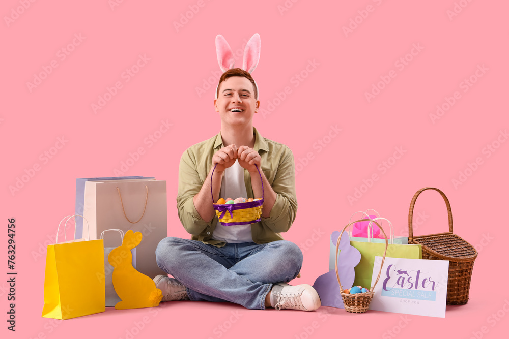 Wall mural Young man in bunny ears with basket and shopping bags sitting on pink background. Easter Sale