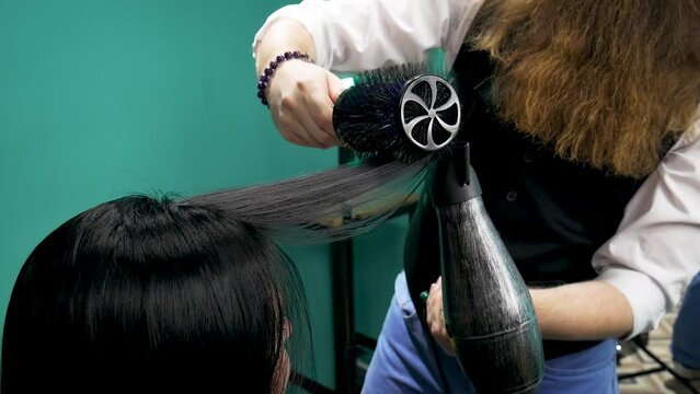 Hairdresser dries woman's hair .Man hairdresser with a long beard in a vest, skillfully does a hairstyle styling with a comb and a hairdryer, to a woman with long black hair in a hairdresser's shop