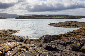 Textured rocks and rich marine flora frame the peaceful expanse of sea leading to rolling hills under a soft sky near Isle of Skye