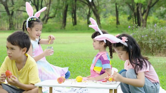 Happy group children in park, cute asian girls and boys with friend, paint egg with paintbrush and blowing soap bubbles  together on green grass in garden. Kids celebrate Easter holiday outdoor