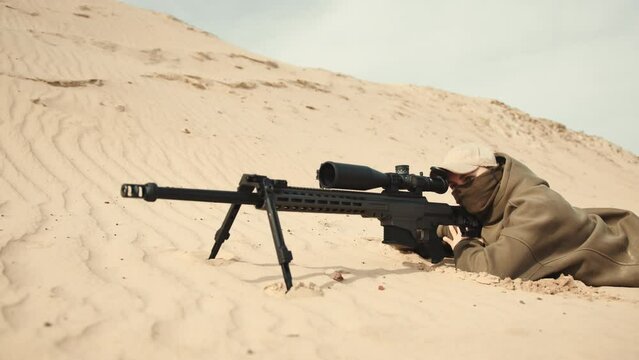 A sniper lies still on sandy ground, blending into the environment while waiting patiently for the perfect moment to take the critical shot. Camera 8K RAW. 