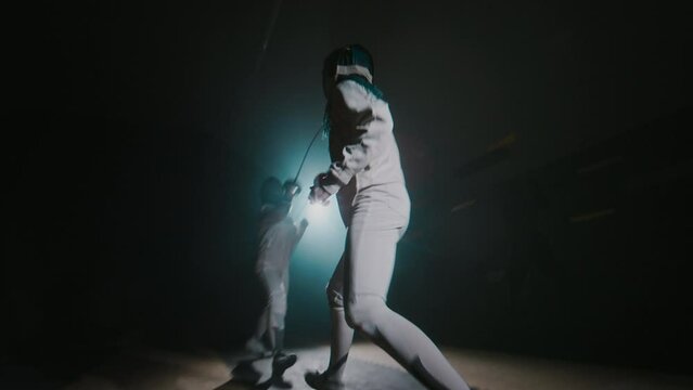 Rear full footage of female foil fencer parrying her partners strikes and riposting while lunging and moving around during tough duel in dark smokey hall