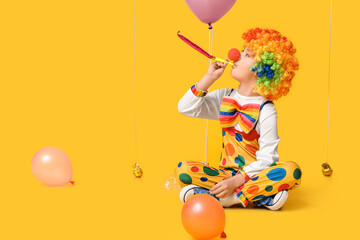 Funny little boy in clown costume with party whistle and balloons sitting on yellow background. April Fools Day celebration - obrazy, fototapety, plakaty