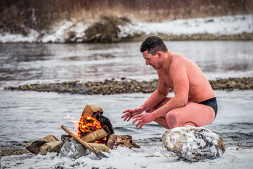 A man walrus warms himself by a fire after swimming in a winter river