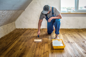Worker in blue overalls laquering the wooden floor with paint roller and eco varnish