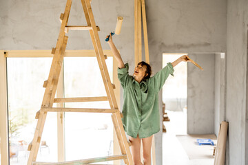 Portrait of a young cute woman standing happily on a ladder with paint roller during repairing process of a house. Creative process of home renovation and repair concept