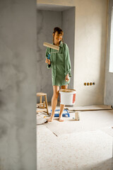Portrait of a young cute woman standing with paint roller and bucket full of paint during repairing process of a house. Creative process of home renovation and repair concept
