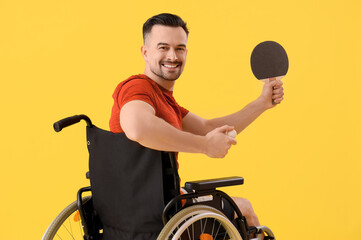 Sporty young man in wheelchair with ping pong racket and ball on yellow background