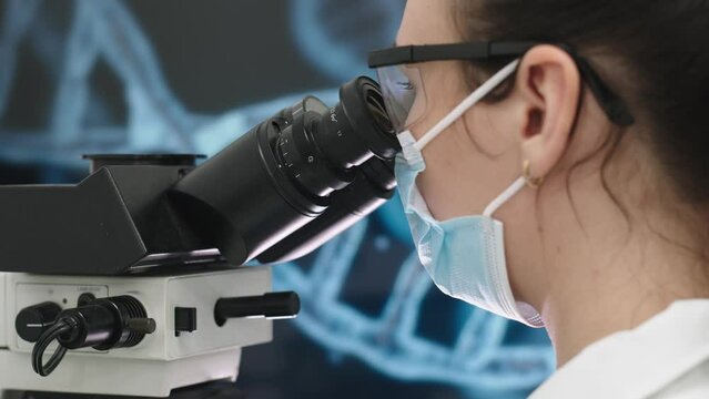 A microbiologist in glasses looks into a microscope, analyzing a sample. Female scientist working with high-tech equipment.