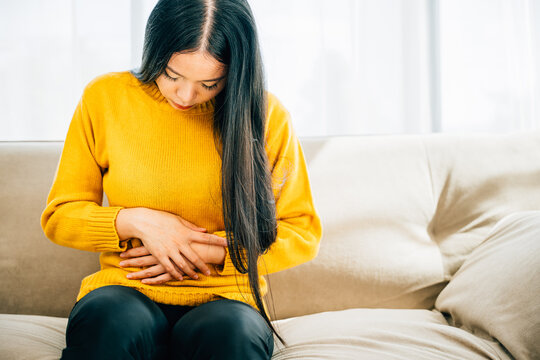 Young woman sitting on a sofa experiences strong abdominal pain at home. Depicting discomfort stomachache and symptoms emphasizing abdominal care. Health care abdomen bloating concept