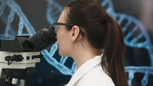 A microbiologist in glasses looks into a microscope, analyzing a sample. Female scientist working with high-tech equipment.