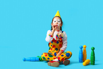 Funny little girl in clown costume with party whistle and juggling clubs on blue background. April Fools' Day celebration