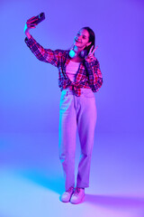 Full-length image of smiling young woman in checkered shirt taking selfie with mobile phone against purple studio background in neon light. Concept of youth, lifestyle, casual fashion, human emotions