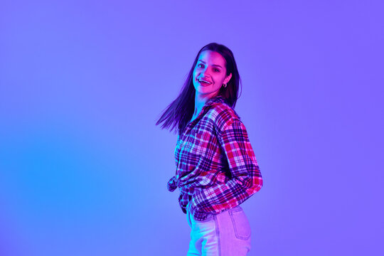 Happy, tender young woman in checkered shirt and jeans dancing and having fun against purple studio background in neon light. Concept of youth, lifestyle, casual fashion, human emotions