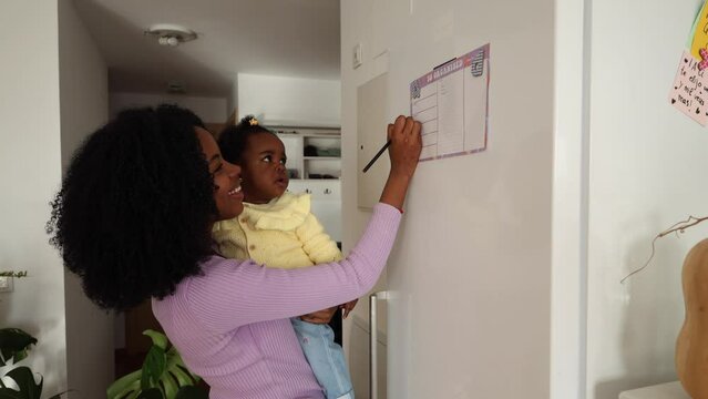 Organized Mother and Baby Planning the Week - Mother holding her baby while writing on a planner attached to the fridge. - Powered by Adobe