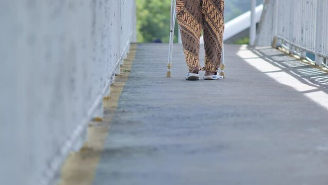 An elderly Asian man with a disabled leg is hobbling using crutches on the Jakarta JPO in the morning