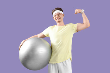 Sporty young man with fitball showing muscles on lilac background