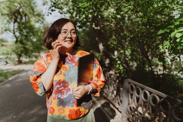 Happy woman chatting on the phone holding a laptop walking in the park Smiling freelancer wearing a colorful blouse using smartphone outdoors